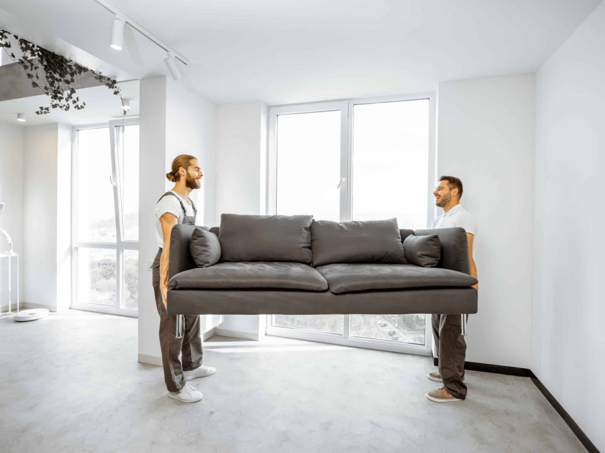 Two professional movers in workwear placing large couch in the living room of the modern white apartment. Furniture delivery concept
